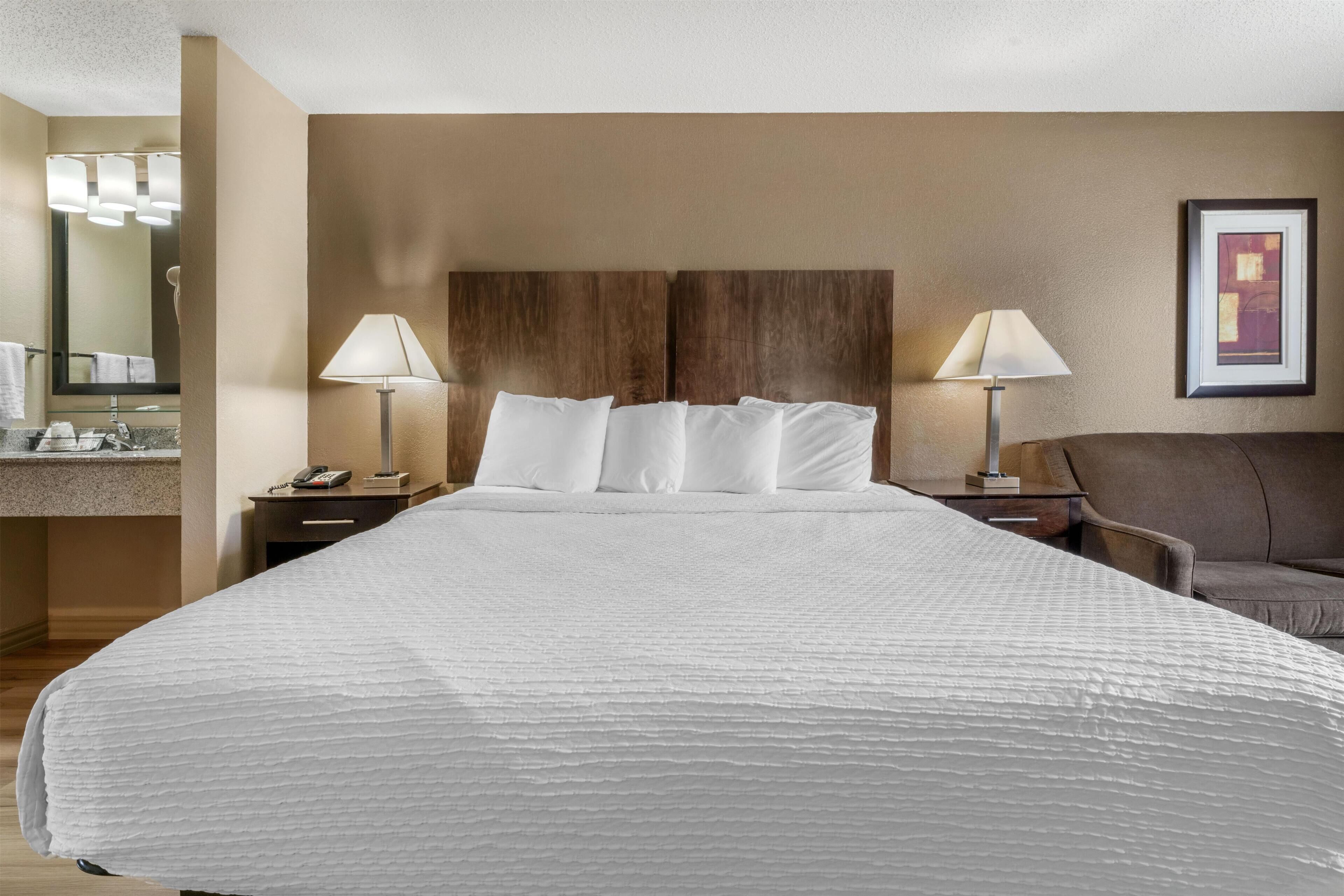 Habitación de hotel con una cama grande, dos lámparas de noche, un cabecero de madera, un baño con espejo y un cuadro en la pared.