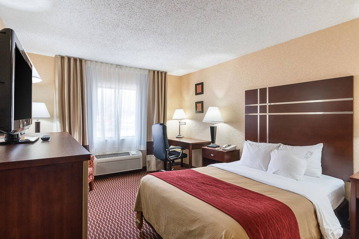 A hotel room with a bed, desk, chair, TV, lamps, and an air conditioning unit.