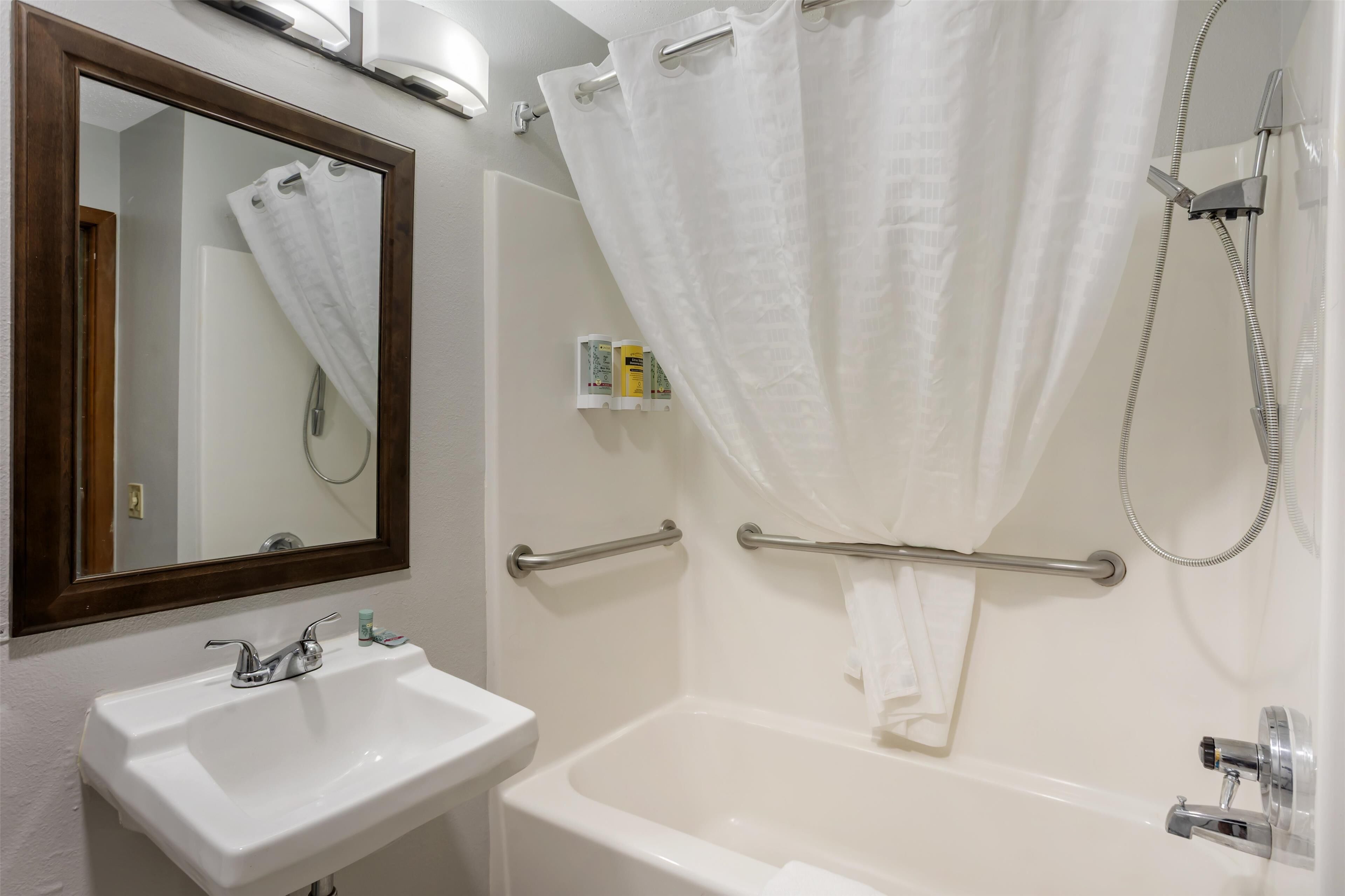 Un baño con lavamanos blanco, espejo y bañera. Hay una cortina de ducha y un perchero.