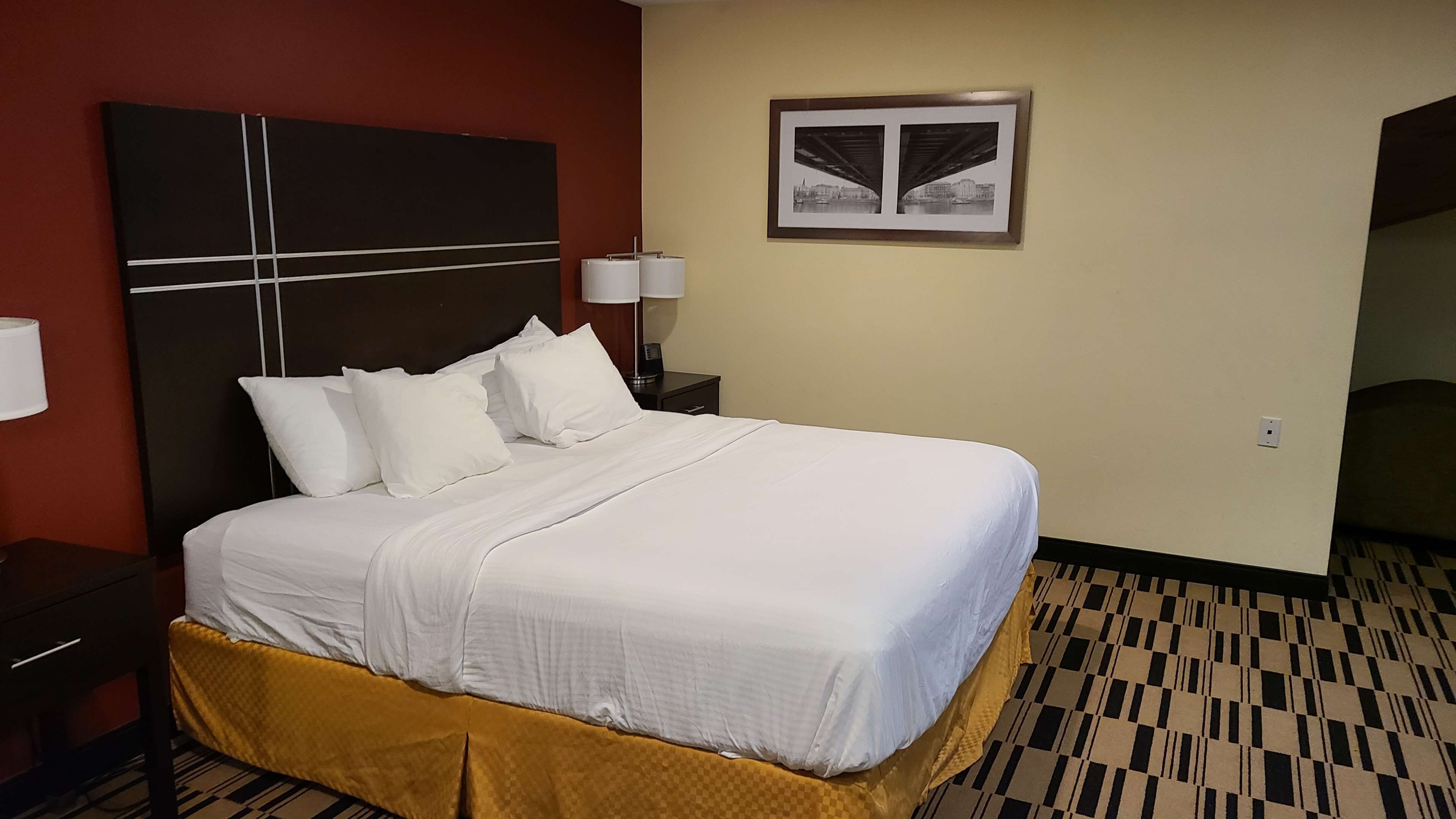 A hotel room with a large bed, two bedside tables, a headboard, a framed picture on the wall, and a patterned carpet.