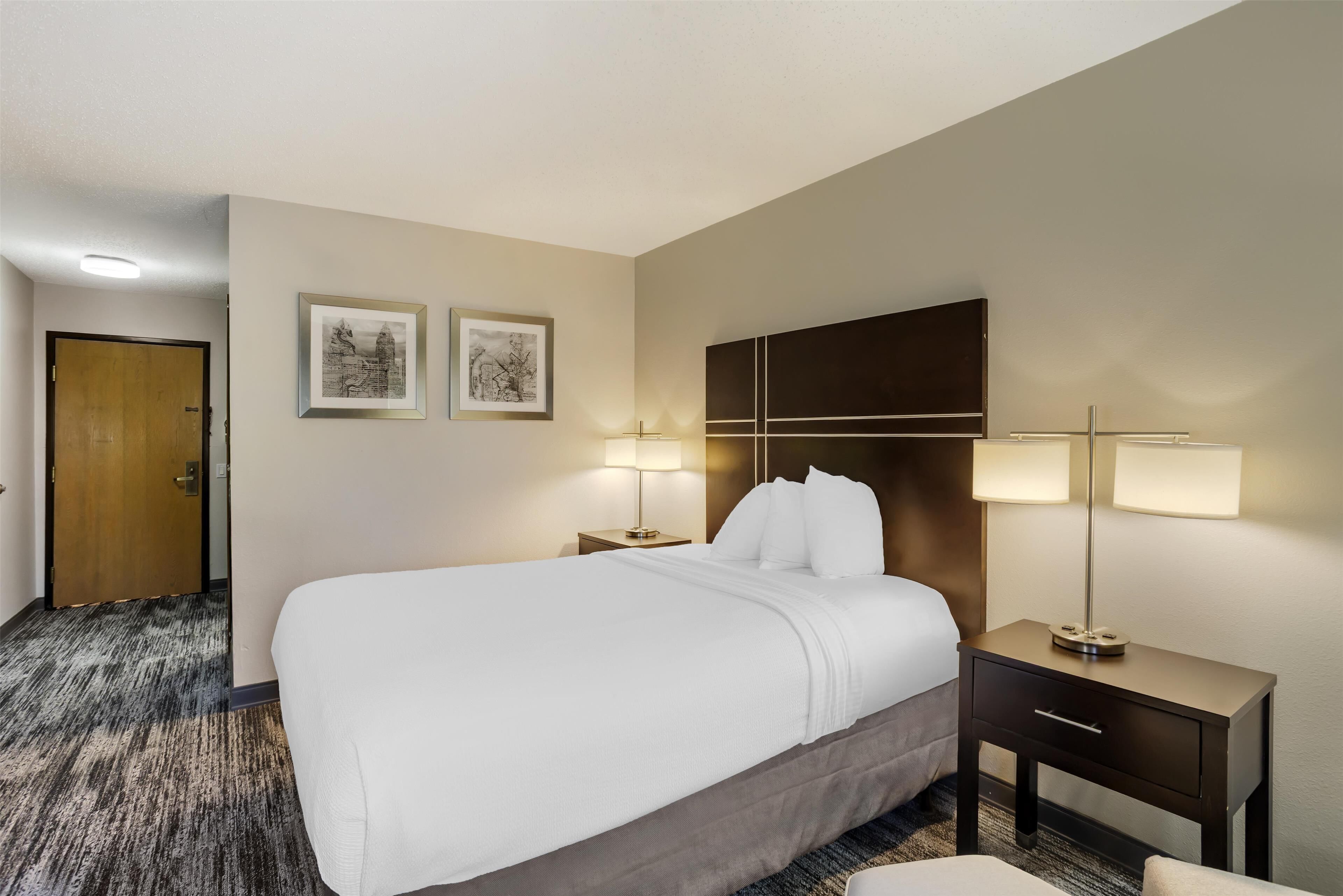 Une chambre d’hôtel avec un grand lit, deux tables de chevet avec des lampes, une porte en bois et deux tableaux encadrés au mur.