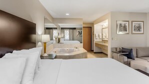 Una habitación de hotel moderna con una cama grande, un baño separado con bañera, una zona de estar con sofá y una mesita.