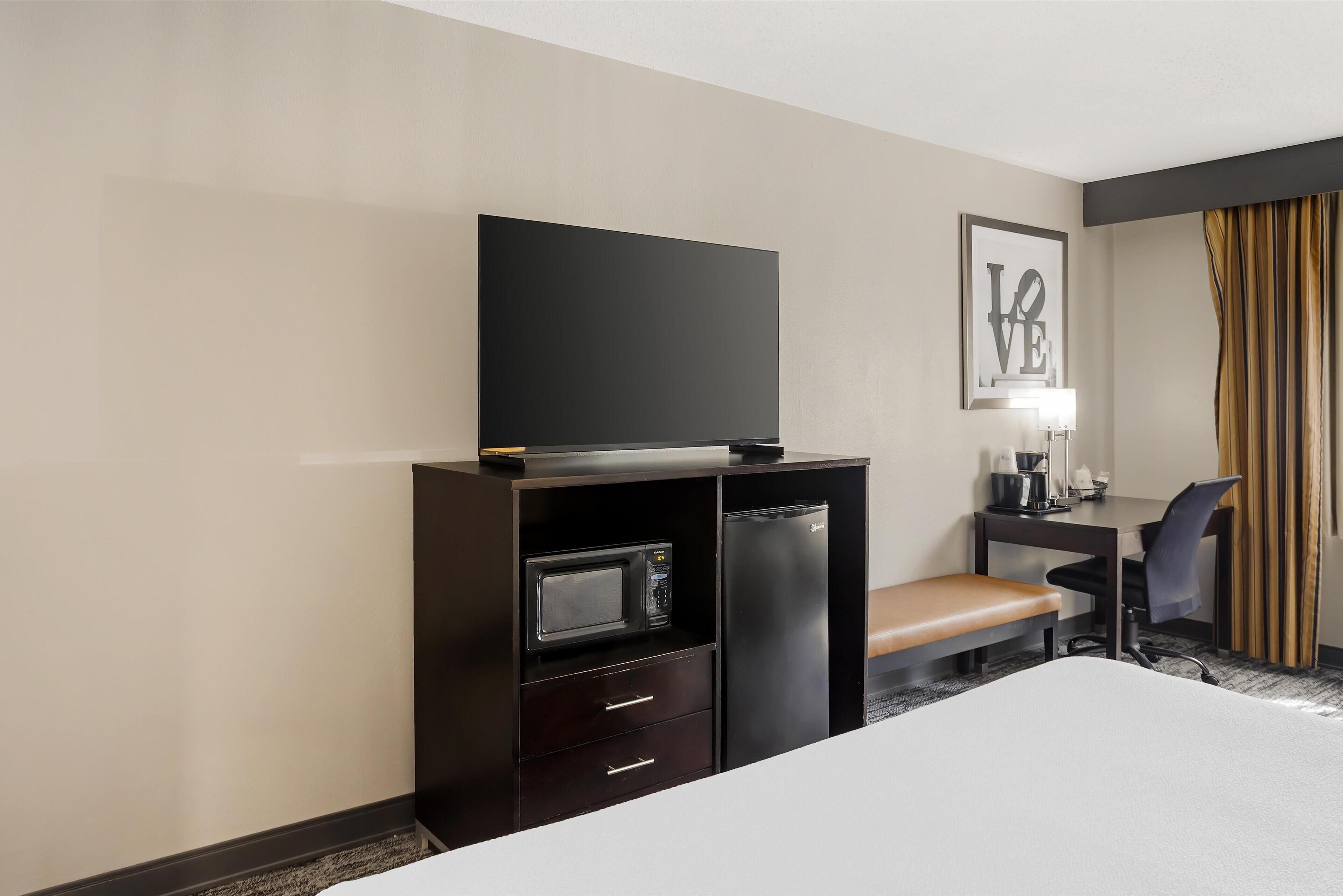A hotel room with a bed, a TV on a cabinet, a microwave, a refrigerator, a desk with a chair, and a bench.