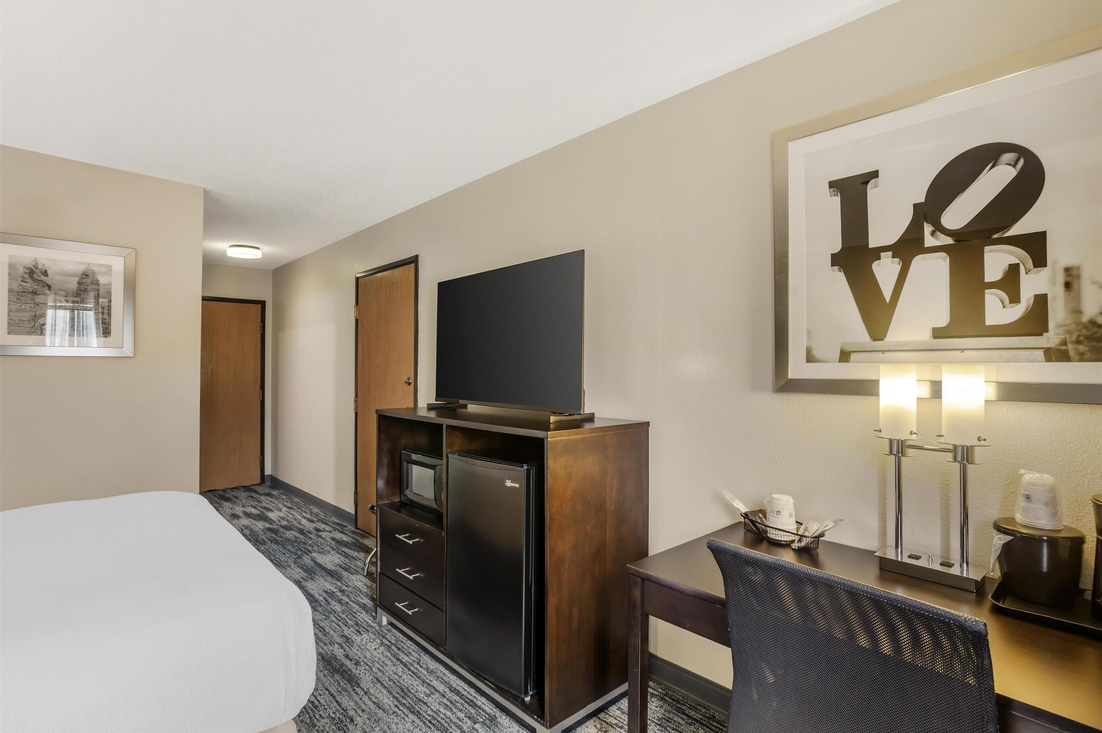 Een hotelkamer met een bed, een televisie op een houten kast, een bureau met een stoel, een koffiezetapparaat en een kunstwerk in een lijst aan de muur.