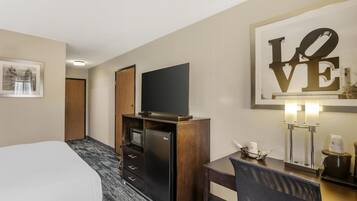 Een hotelkamer met een bed, een televisie op een houten kast, een bureau met een stoel, een koffiezetapparaat en een kunstwerk in een lijst aan de muur.