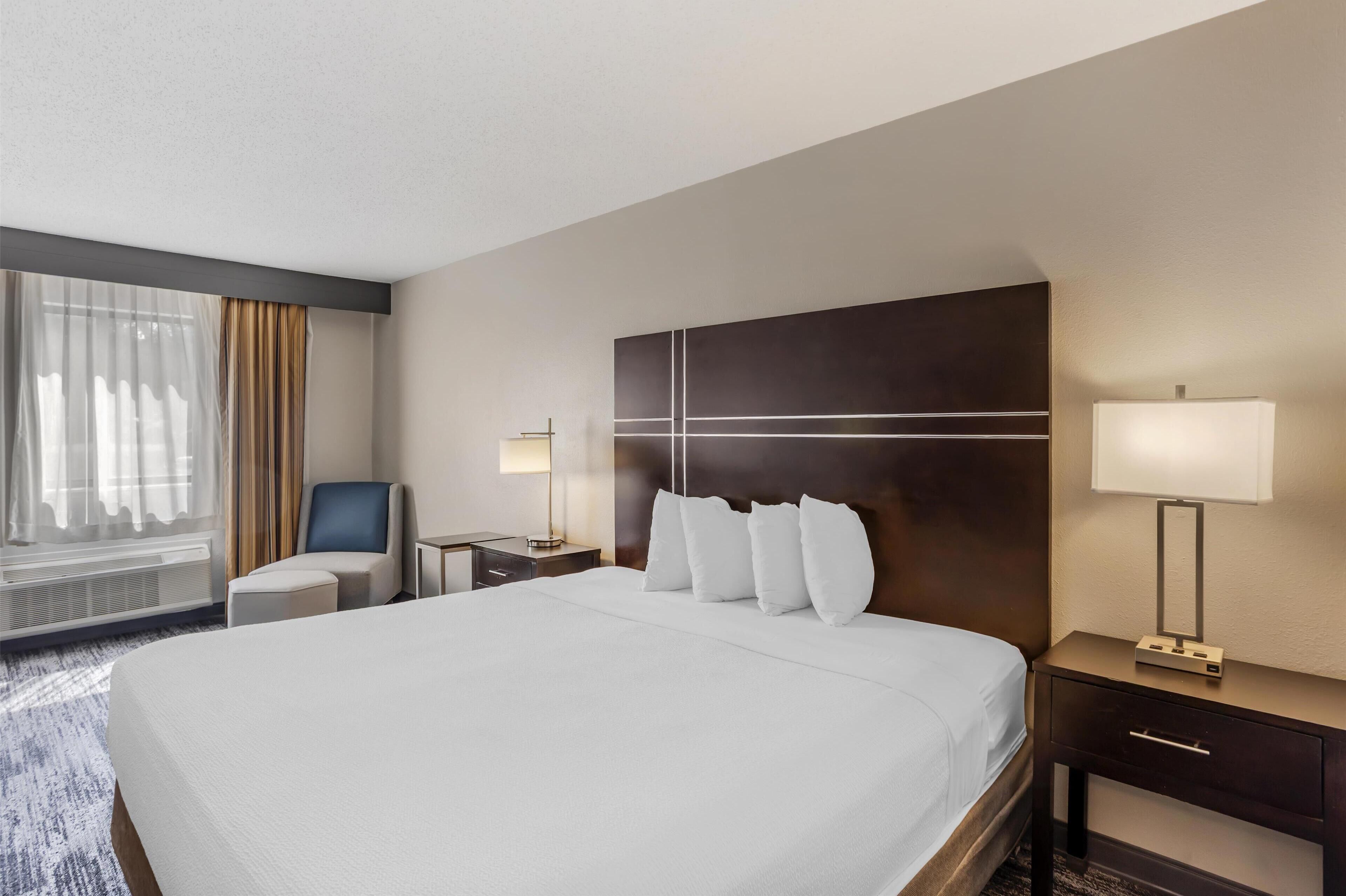 Een hotelkamer met een groot bed, nachtkastje met lamp, een stoel en een raam met gordijnen.