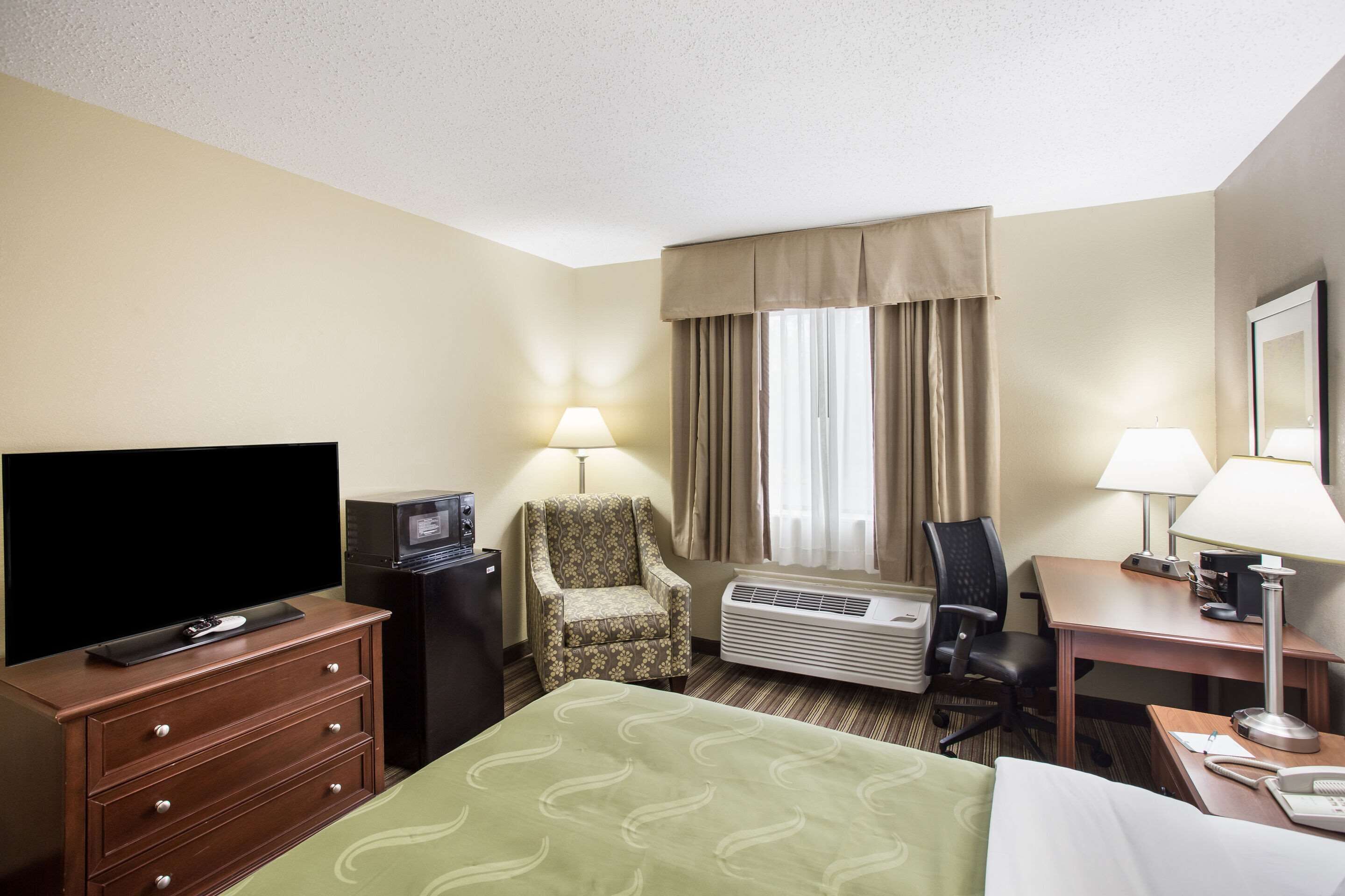 A hotel room with a bed, a desk, a chair, a television, and an air conditioning unit.