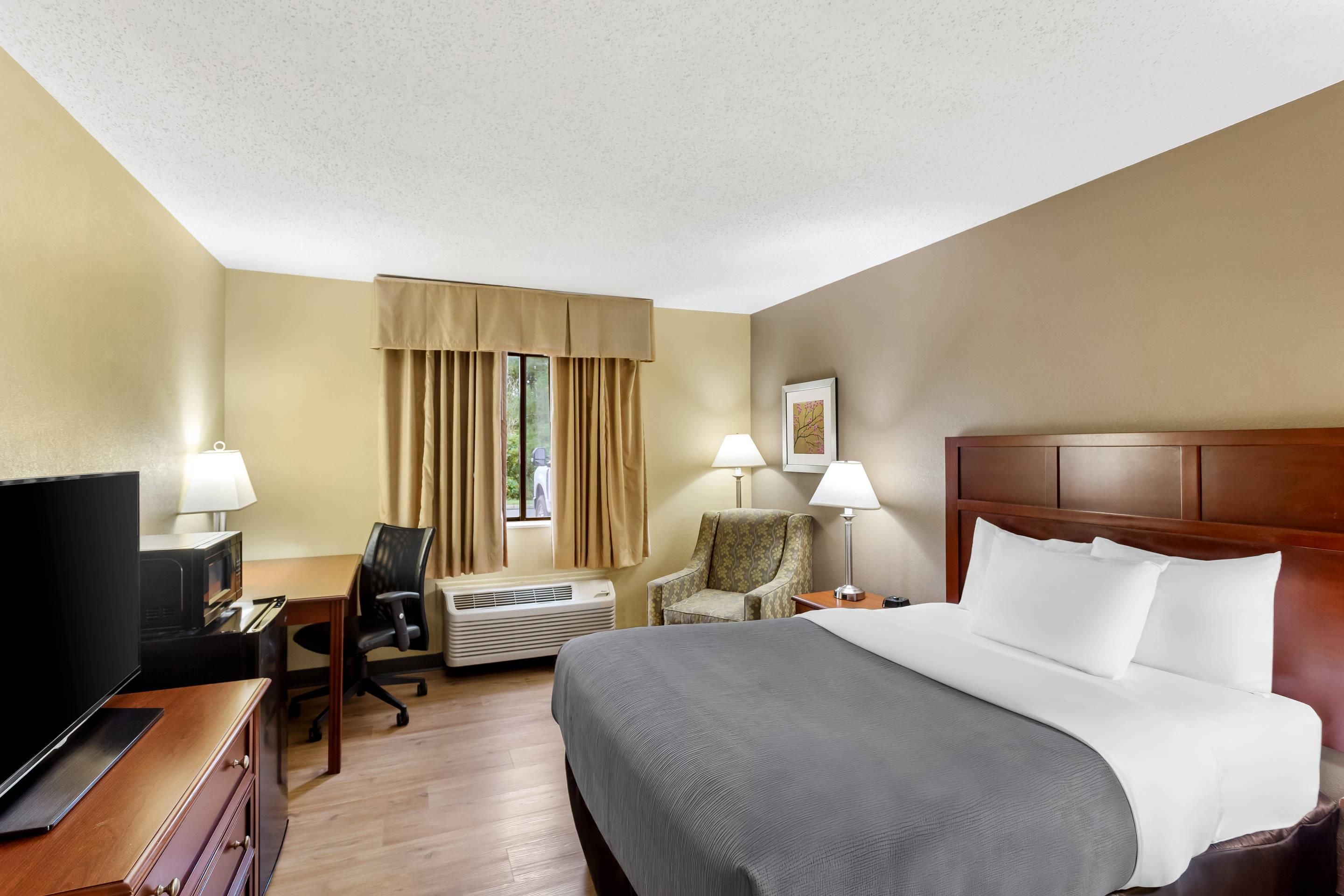 Quarto de hotel com cama, escrivaninha, cadeira, televisão e janela com cortinas.