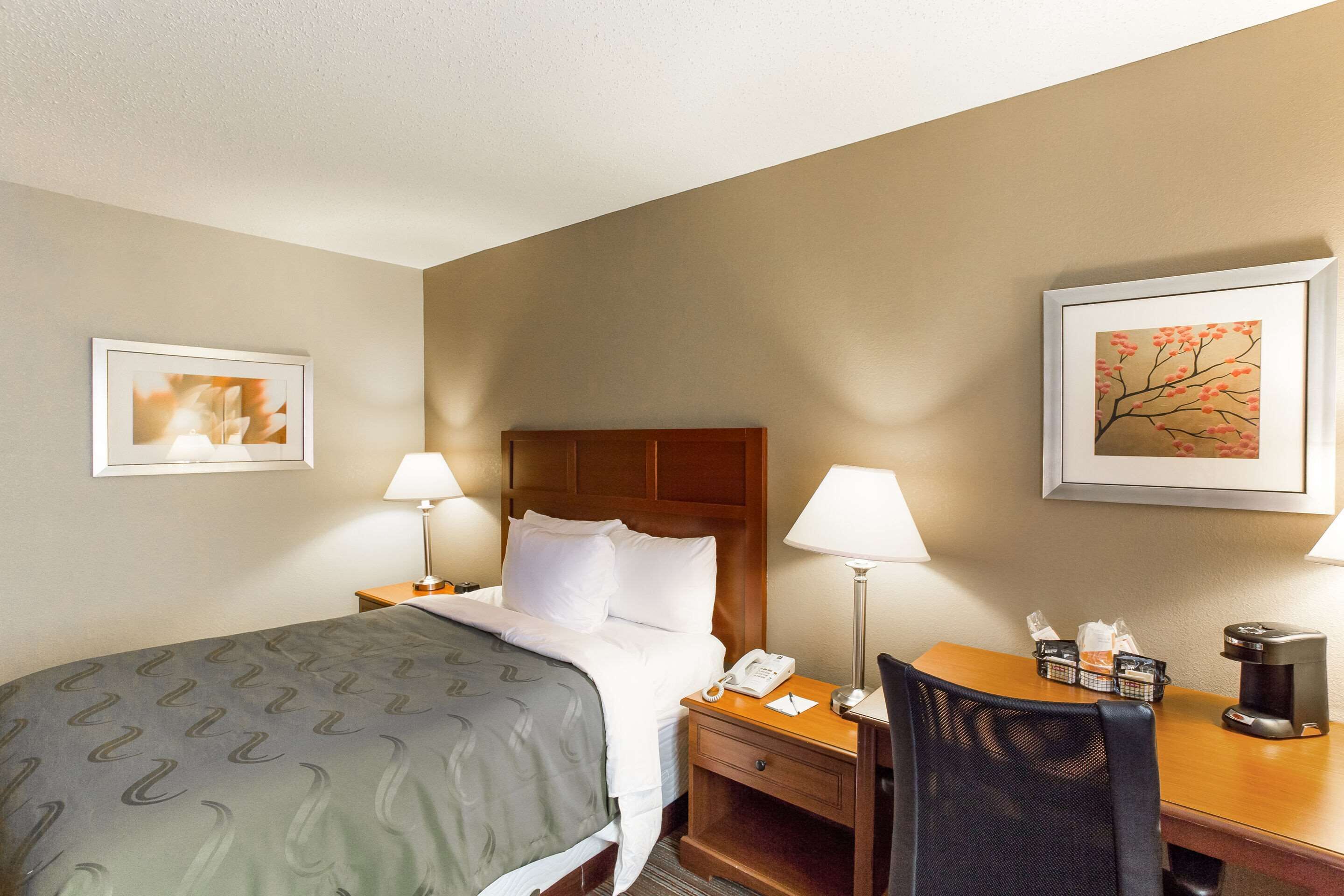 A hotel room with a bed, bedside tables, a desk with a chair, a coffee maker, and framed artwork on the walls.