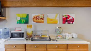 A breakfast bar with a microwave, waffle maker, and fruit display.