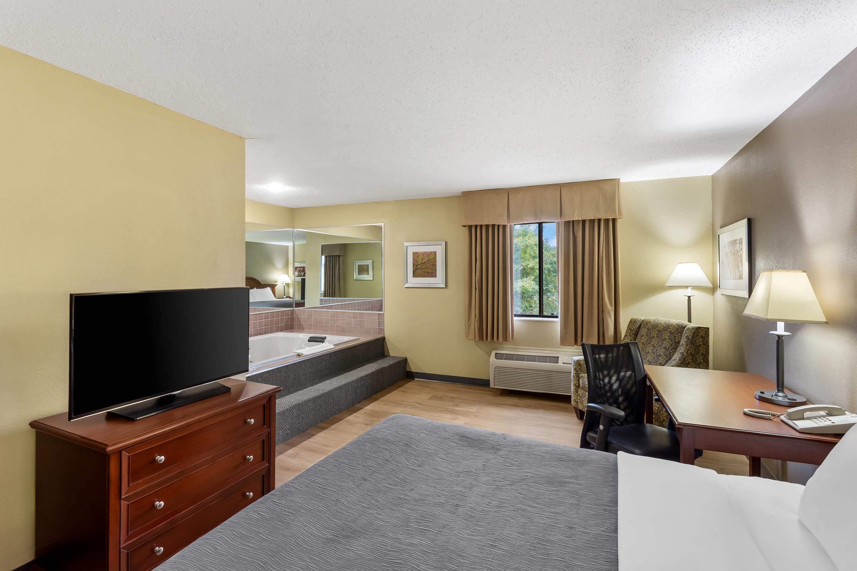 A hotel room with a television, a desk with a lamp, a chair, a bed, a window with curtains, and a bathroom with a bathtub.