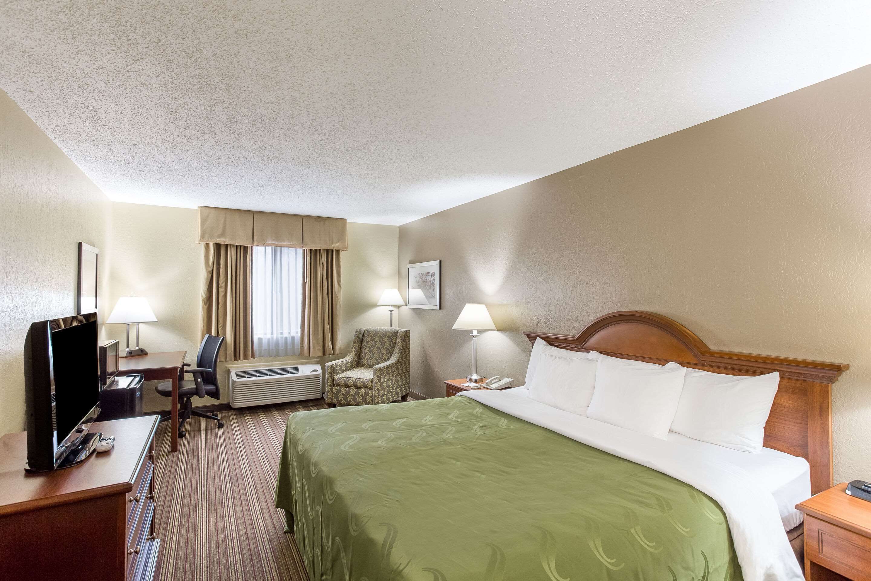A hotel room with a bed, a desk with a television, a chair, a lamp, and a window with curtains.