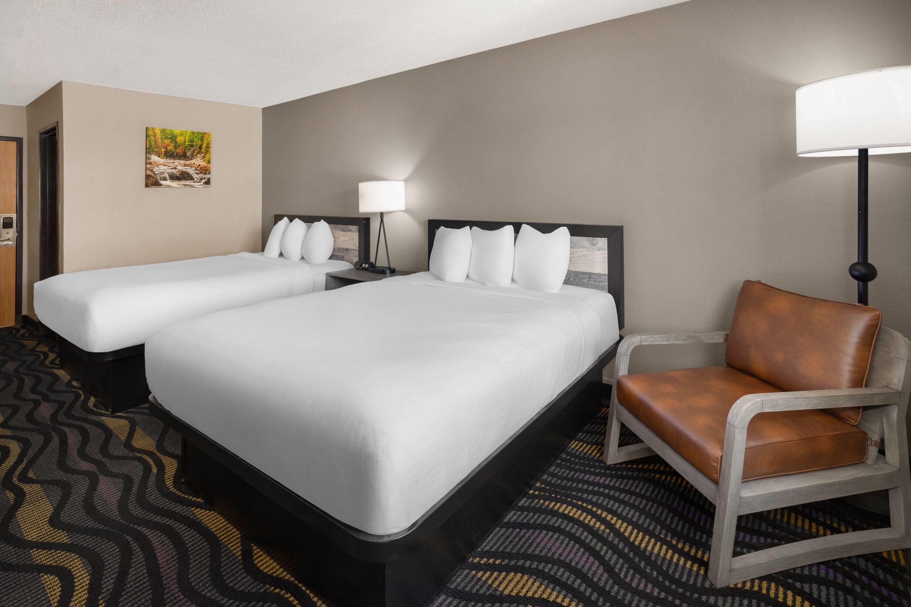 A hotel room with two beds, a chair, a lamp, and a painting on the wall.