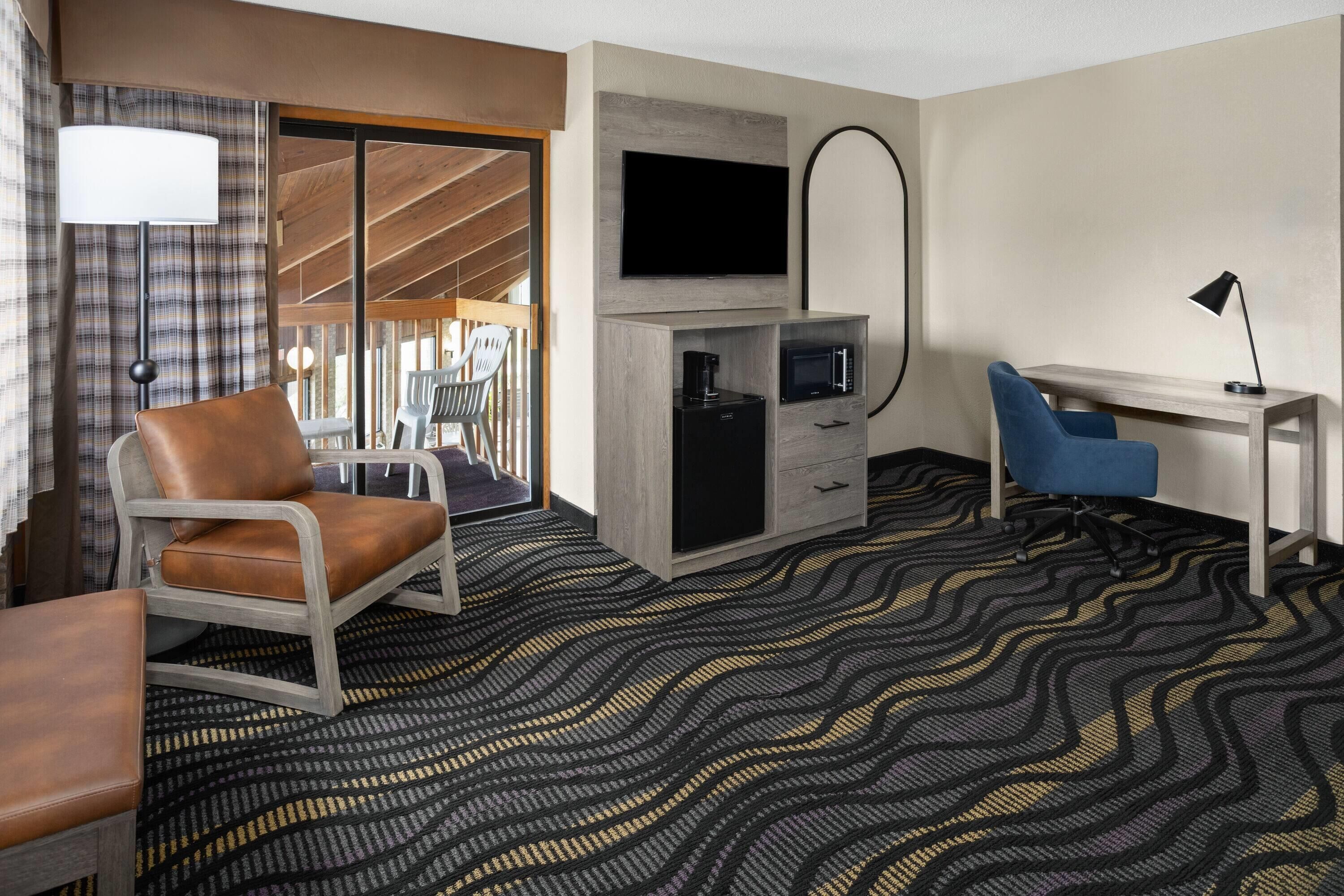 A hotel room with a brown leather chair, a desk with a computer, a flat-screen TV, a minibar, and a patterned carpet.