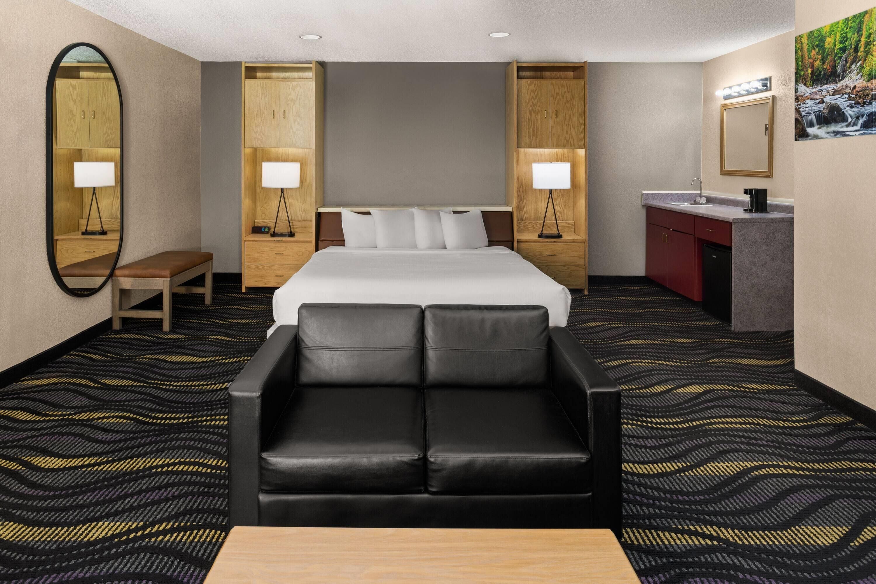 A hotel room with a large bed, a sofa, a desk, a mirror, a nightstand with a lamp, a vanity with a mirror, and a painting on the wall.