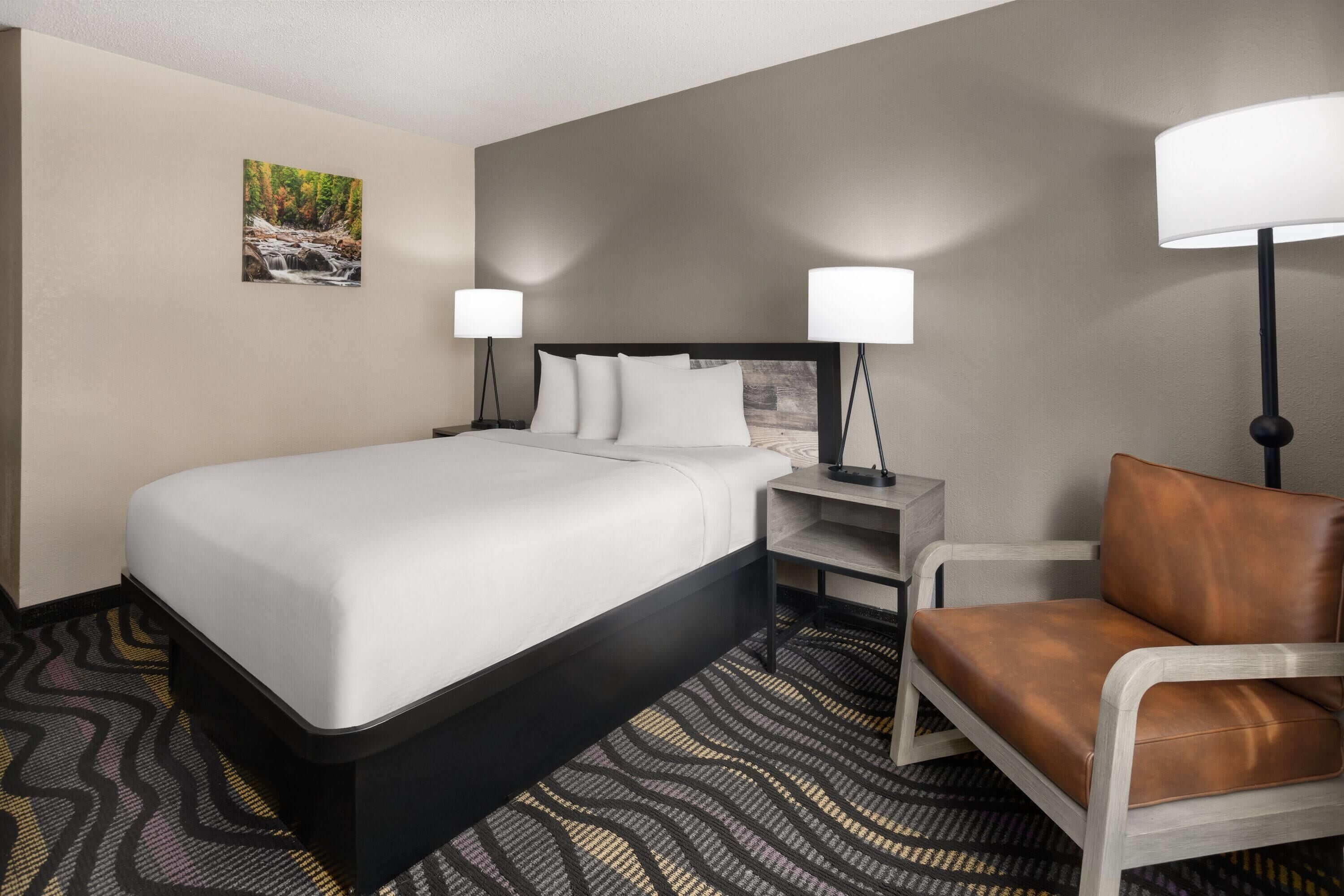 Habitación de hotel con cama, dos lámparas de noche, una silla, una mesita de noche y un cuadro en la pared.