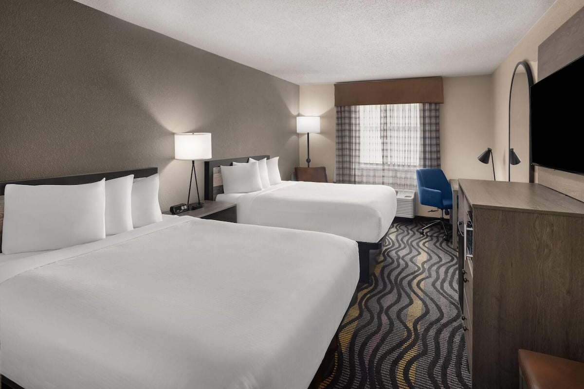 A hotel room with two beds, a desk, a chair, a TV, and a lamp.