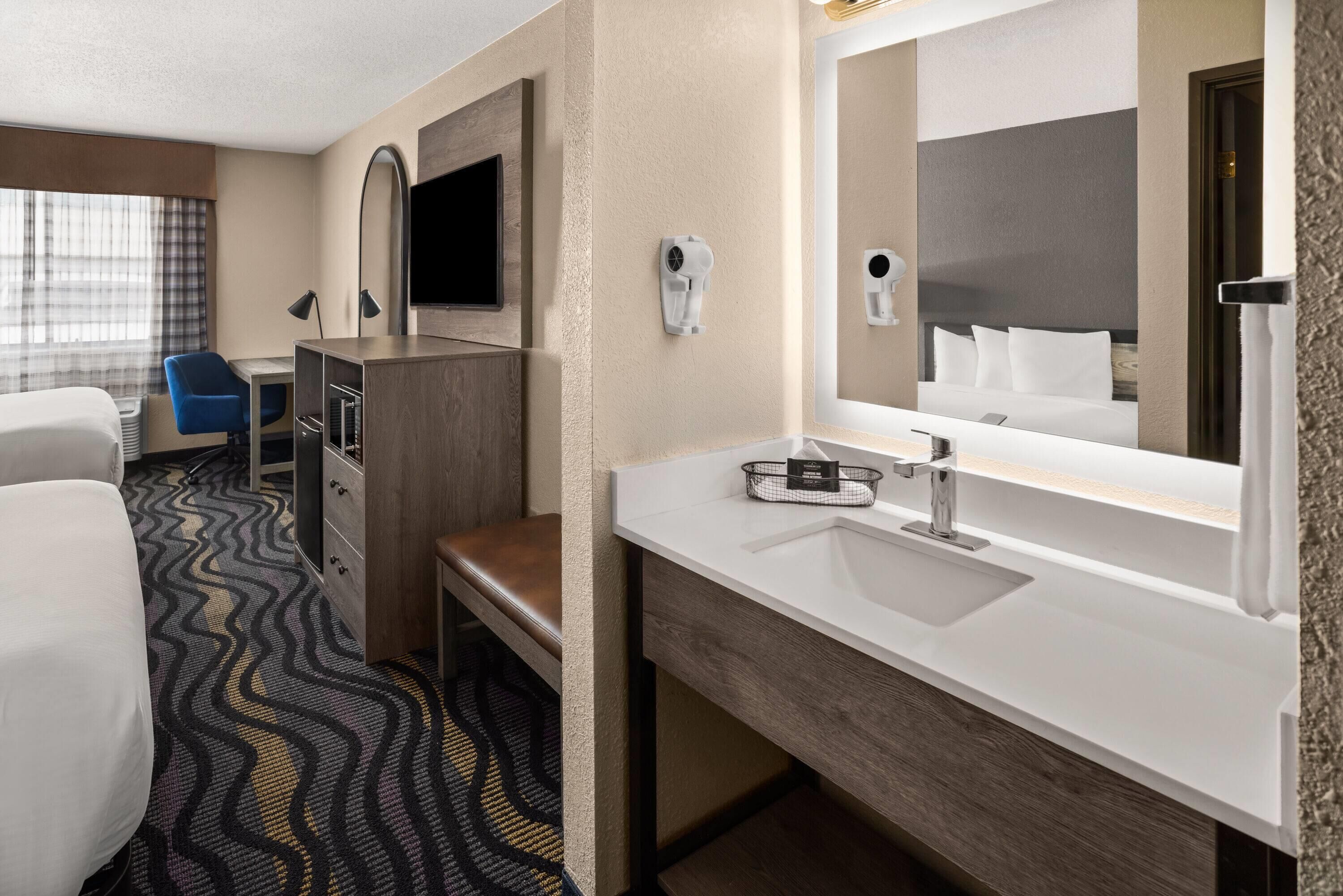A hotel room with a bed, a desk with a chair, a large mirror, a sink, and a hairdryer.