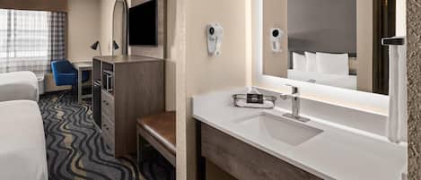 Habitación de hotel con cama, escritorio con silla, espejo grande, lavamanos y secador de pelo.