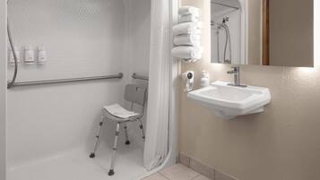 A bathroom with a shower, a wheelchair-accessible chair, a sink, and a mirror.