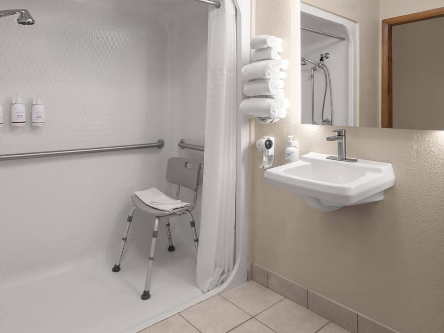 A bathroom with a shower, a wheelchair-accessible chair, a sink, and a mirror.