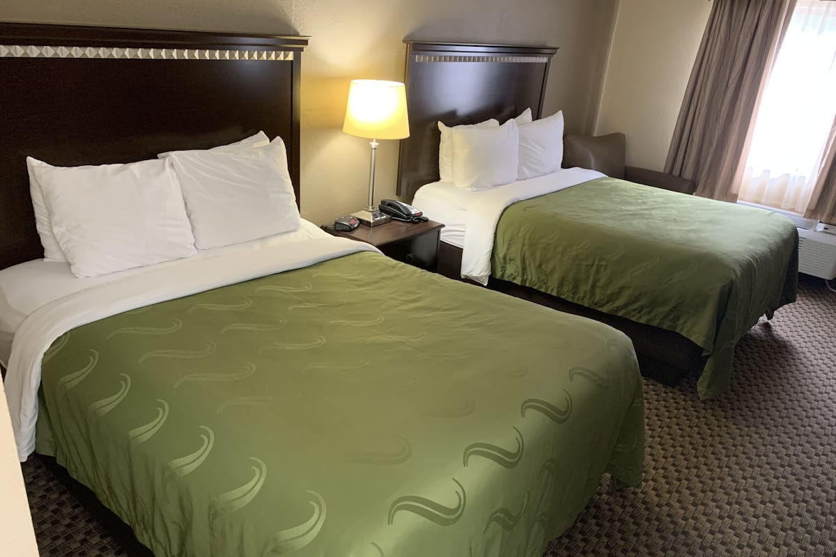 Quarto de hotel com duas camas, cada uma com colcha verde e almofadas brancas. Há uma mesa de cabeceira com abajur entre as camas, e uma janela com cortinas à direita.