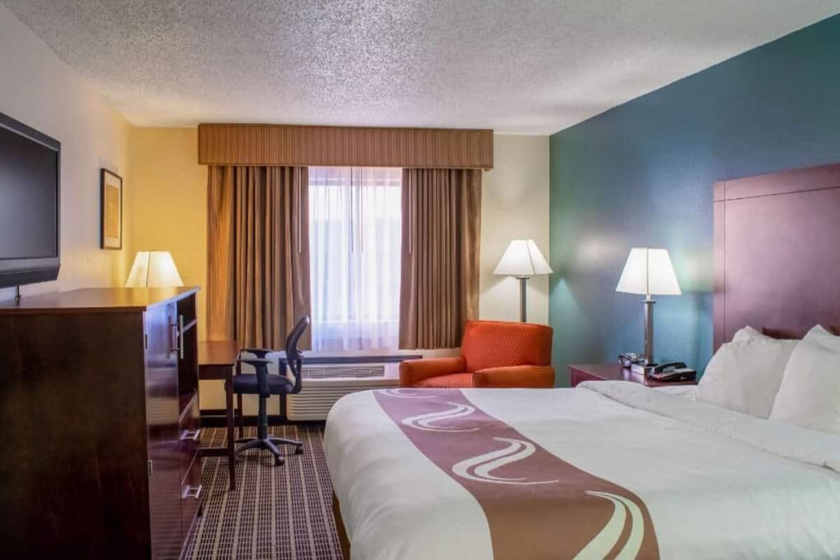 Quarto de hotel com cama, escrivaninha, cadeira, duas luminárias e televisão.