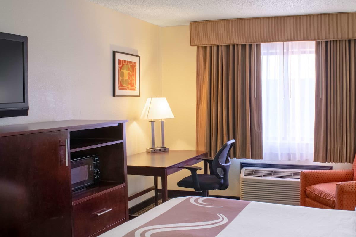 Quarto de hotel com cama, escrivaninha, cadeira, televisão, micro-ondas e janela com cortinas.