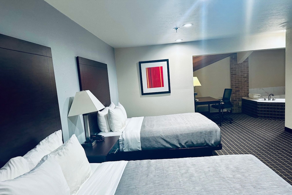 Quarto de hotel com duas camas, uma mesa, uma cadeira, uma luminária de mesa, um quadro emoldurado, um banheiro com banheira e uma parede de tijolos.