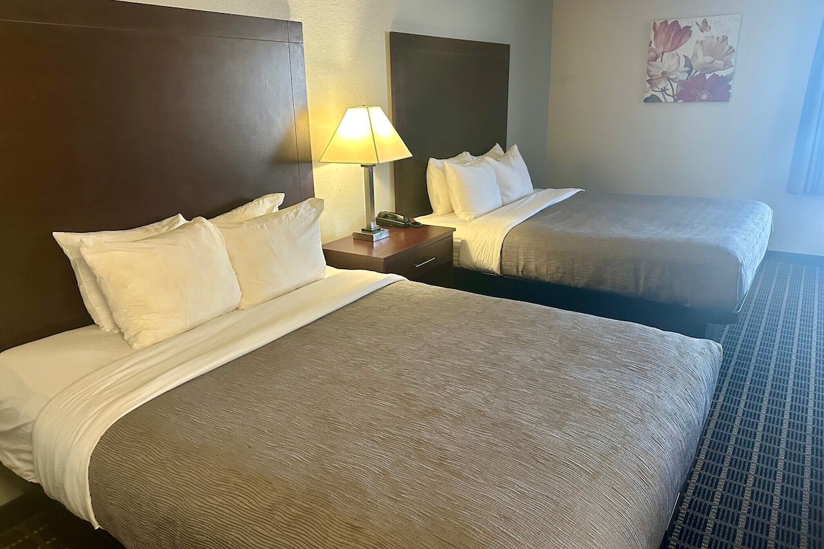 Quarto de hotel com duas camas, uma mesinha de cabeceira com abajur e uma obra de arte floral na parede.
