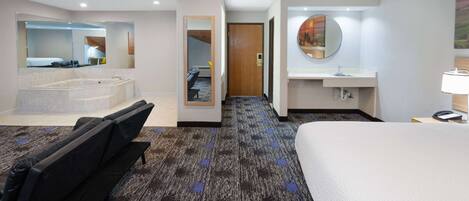 A hotel room with a large bed, a separate seating area with a chair, a bathtub, a large mirror, a sink, and a door leading to another room.
