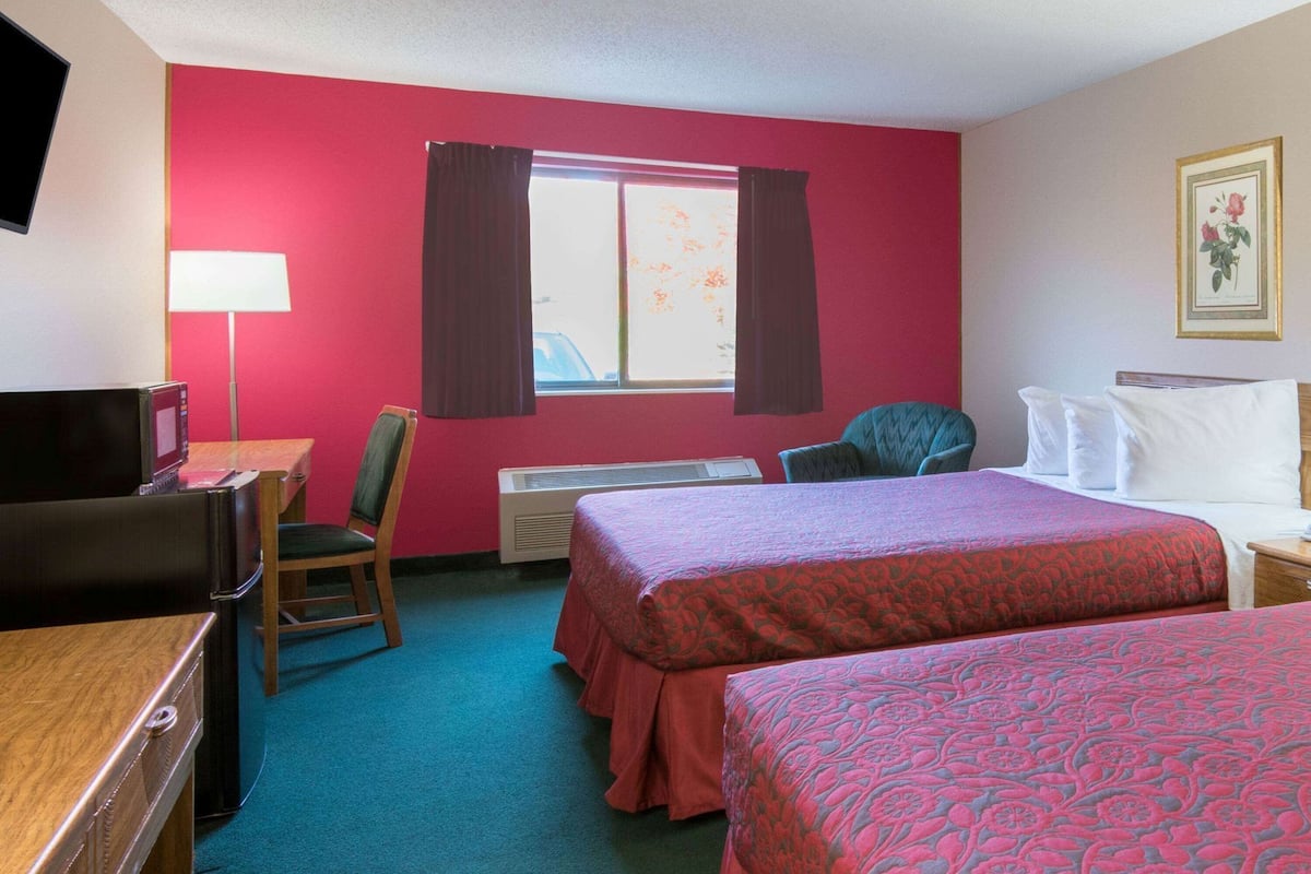 A hotel room with two beds, a desk with a computer, a chair, a window with curtains, and a framed picture on the wall.