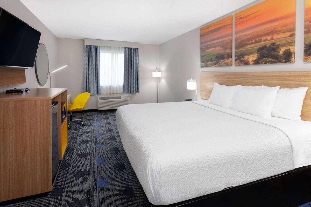 A hotel room with a bed, a desk with a chair, a flat-screen TV, a window with curtains, and a painting on the wall.