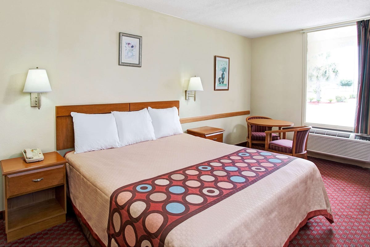 Una habitación de hotel con una cama grande, dos mesitas de noche, una mesita pequeña, dos sillas, una ventana con cortinas y dos lámparas de pared.