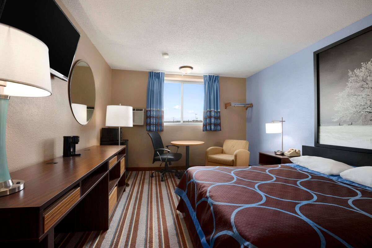 A hotel room with a bed, a desk, a chair, a small table, a lamp, a mirror, and a window with curtains.