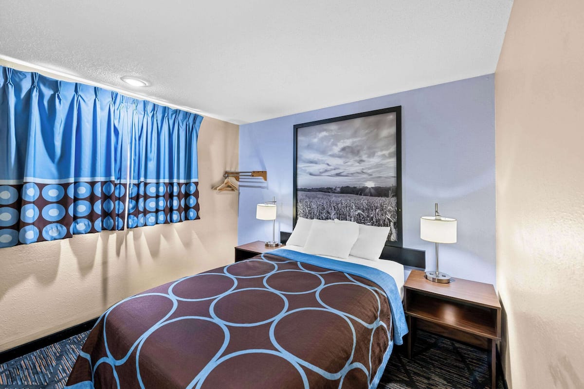 A hotel room with a large bed, two bedside tables with lamps, a window with curtains, and a framed picture on the wall.