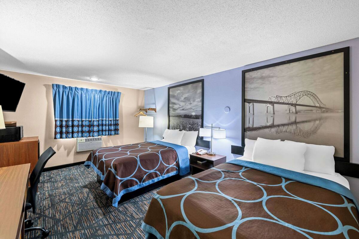 A hotel room with two beds, a desk, a chair, a window with curtains, a wall-mounted TV, and a large framed picture of a bridge.