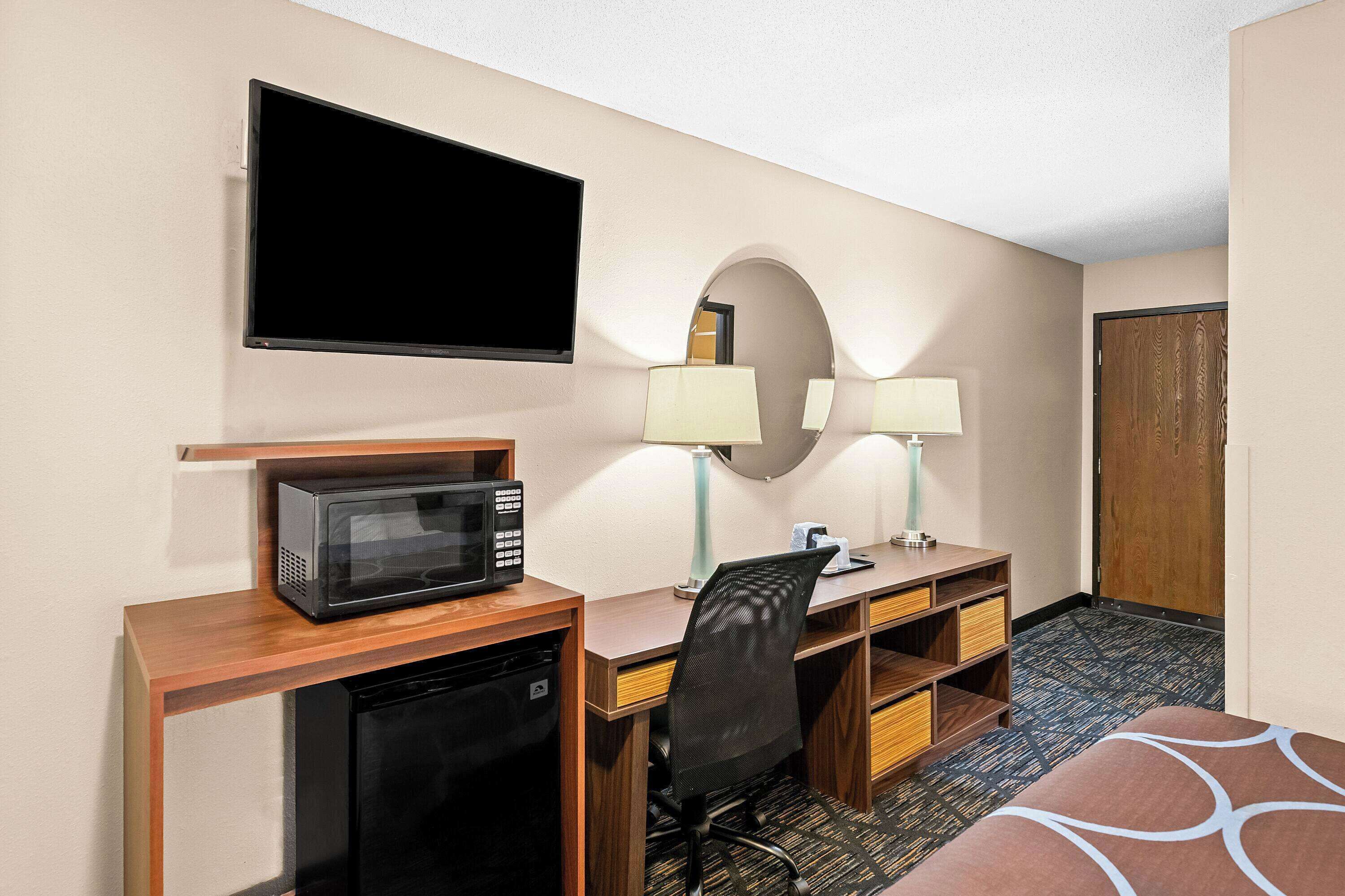 A hotel room with a desk, microwave, chair, lamps, and a TV.