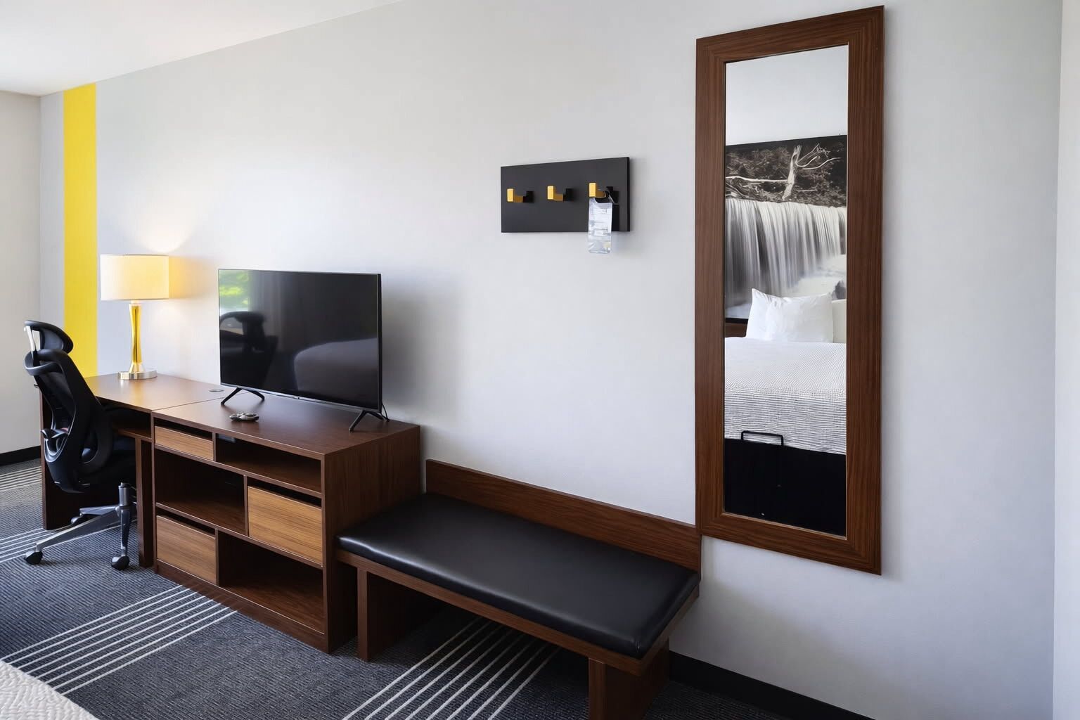A hotel room with a desk, chair, TV, bench, and a large mirror.