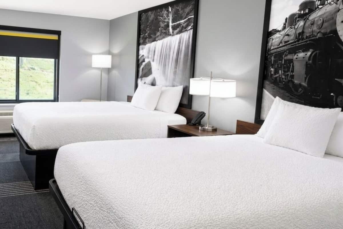 A hotel room with two beds, a desk, a chair, a window with a view, and a large framed picture on the wall.