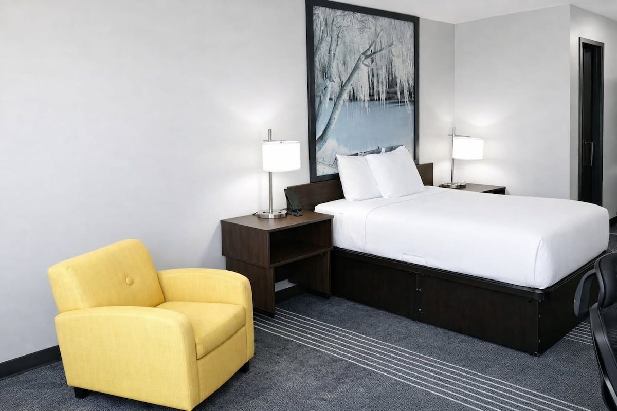 A hotel room with a bed, two bedside tables, a desk, a yellow armchair, and a large window with a view of trees.
