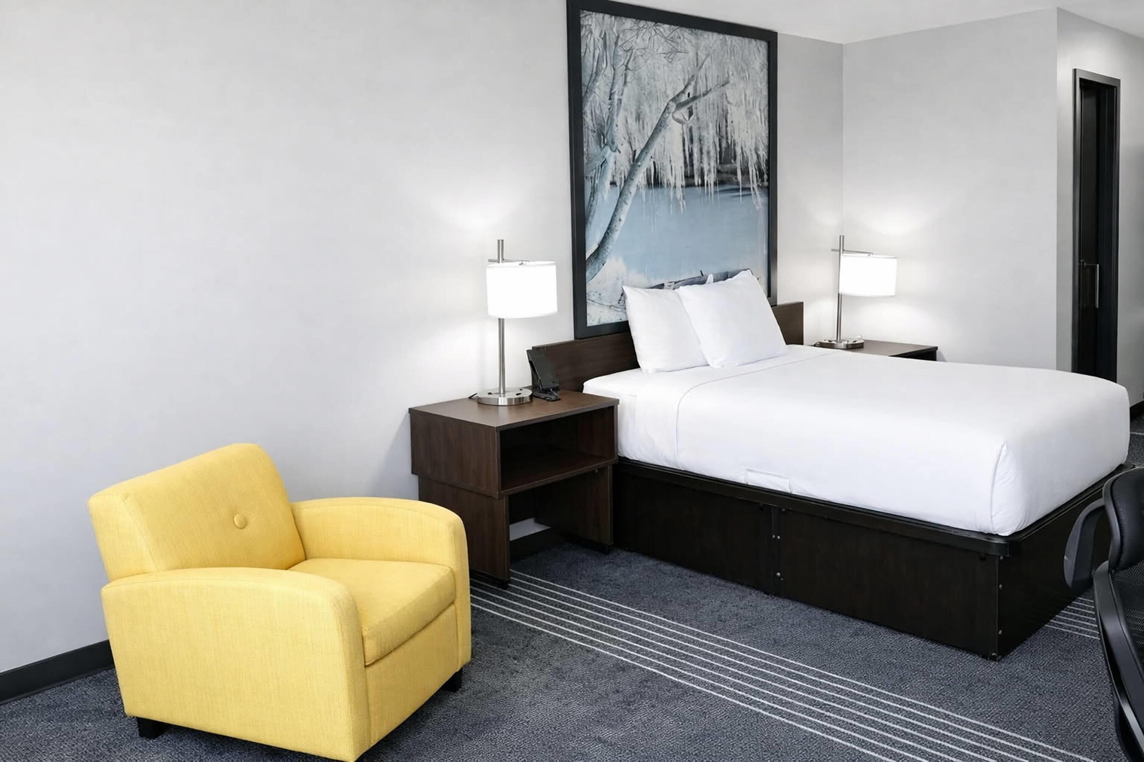 Una habitación de hotel con una cama, dos lámparas de noche, una mesita de noche, un sillón amarillo y un cuadro grande de un paisaje invernal.