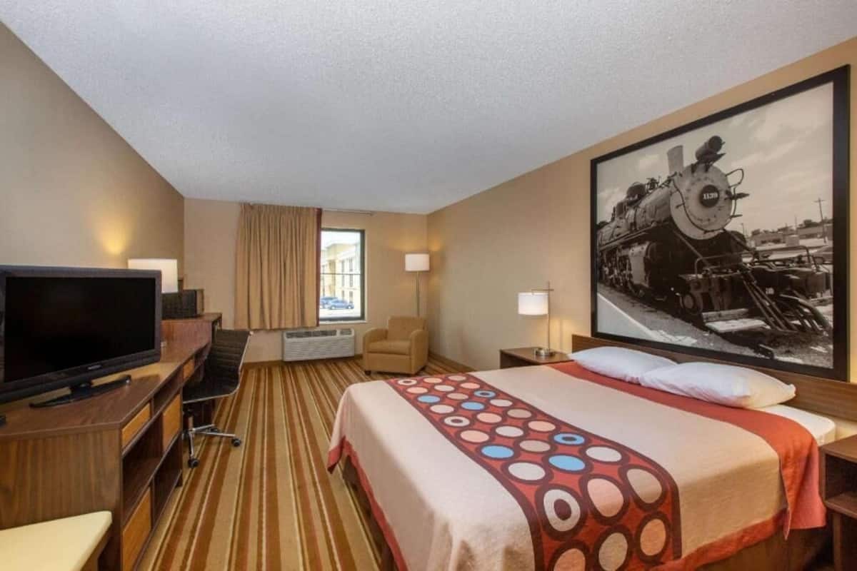 A hotel room with a bed, a television, a desk, a chair, a lamp, and a large framed picture of a train.