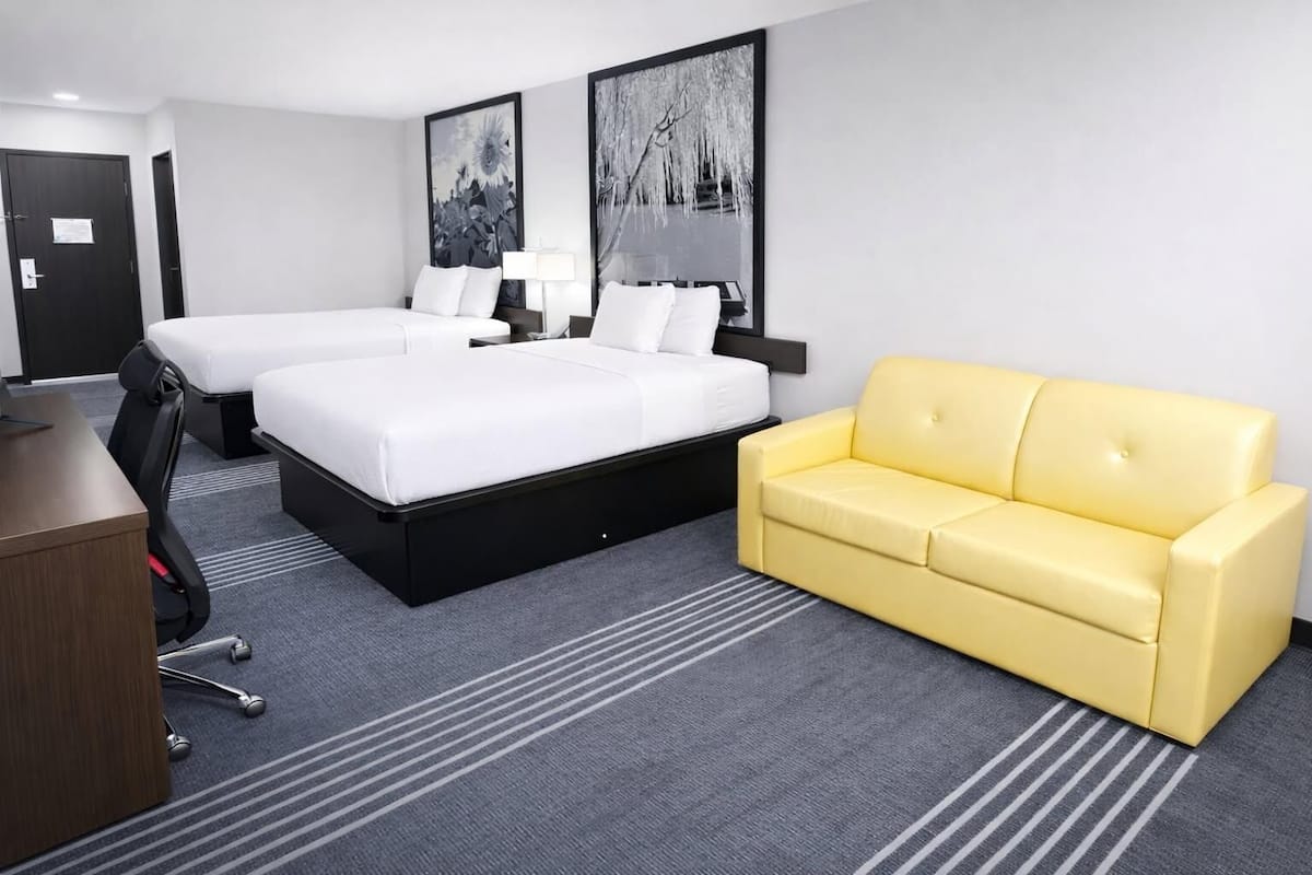 A hotel room with two beds, a yellow sofa, a desk, and two framed pictures on the wall.