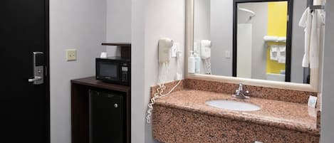 Un bagno d'albergo con lavandino, specchio, porta asciugamani, forno a microonde e un piccolo frigorifero.