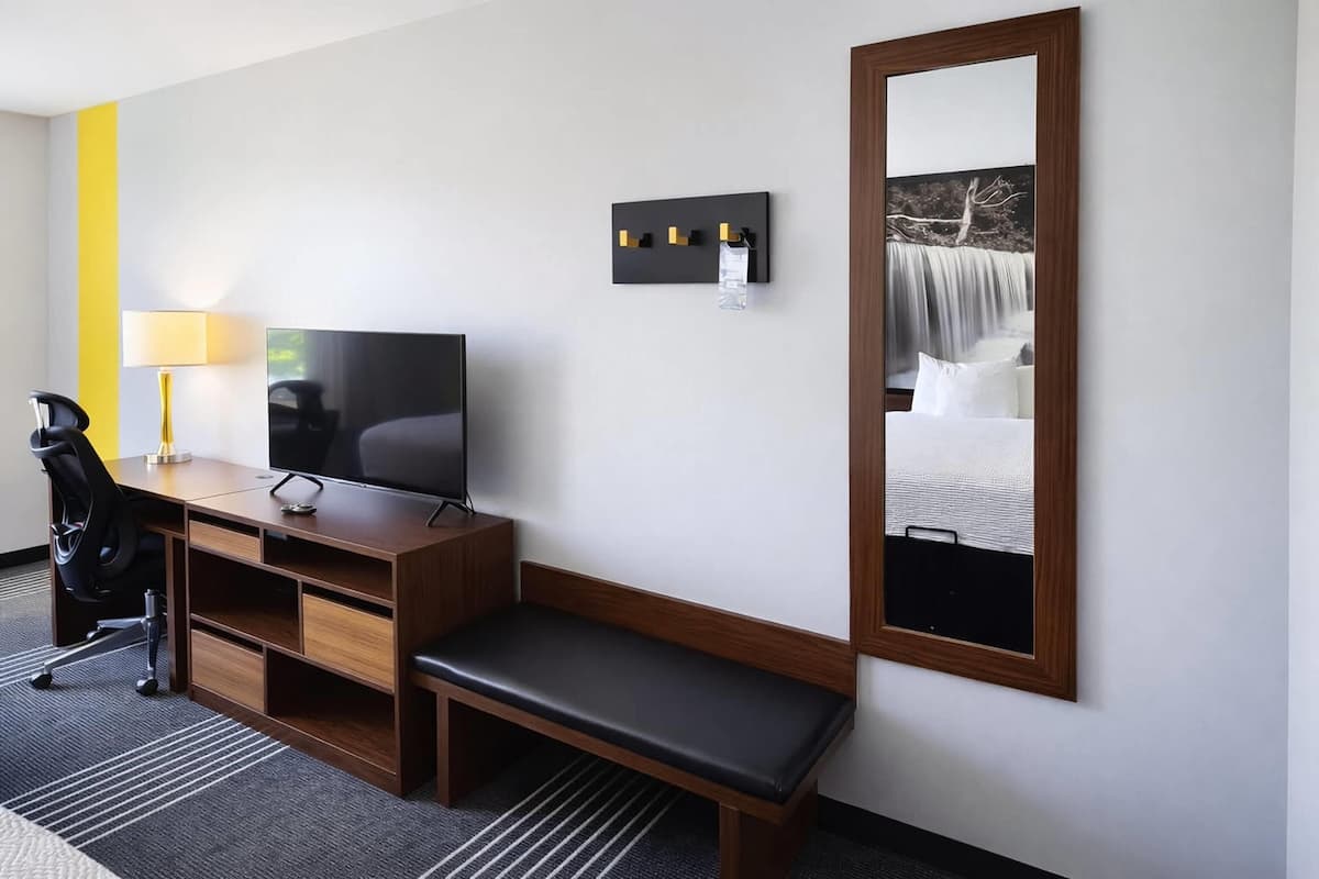 A hotel room with a wooden desk, a television, a bench, a mirror, and a wall-mounted light fixture.