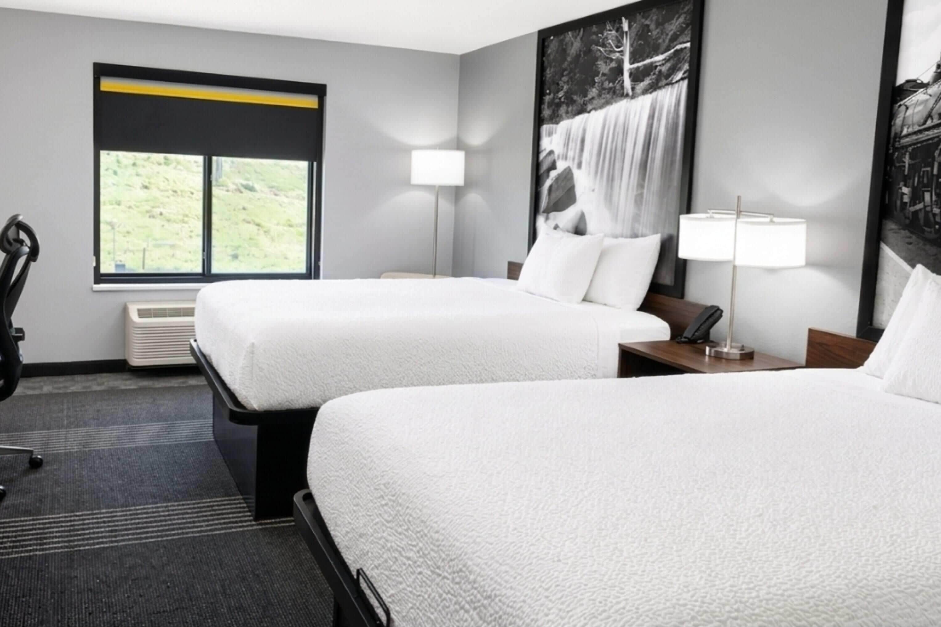 Habitación de hotel con dos camas, un escritorio, una silla, una ventana con vistas y una lámpara de pared.