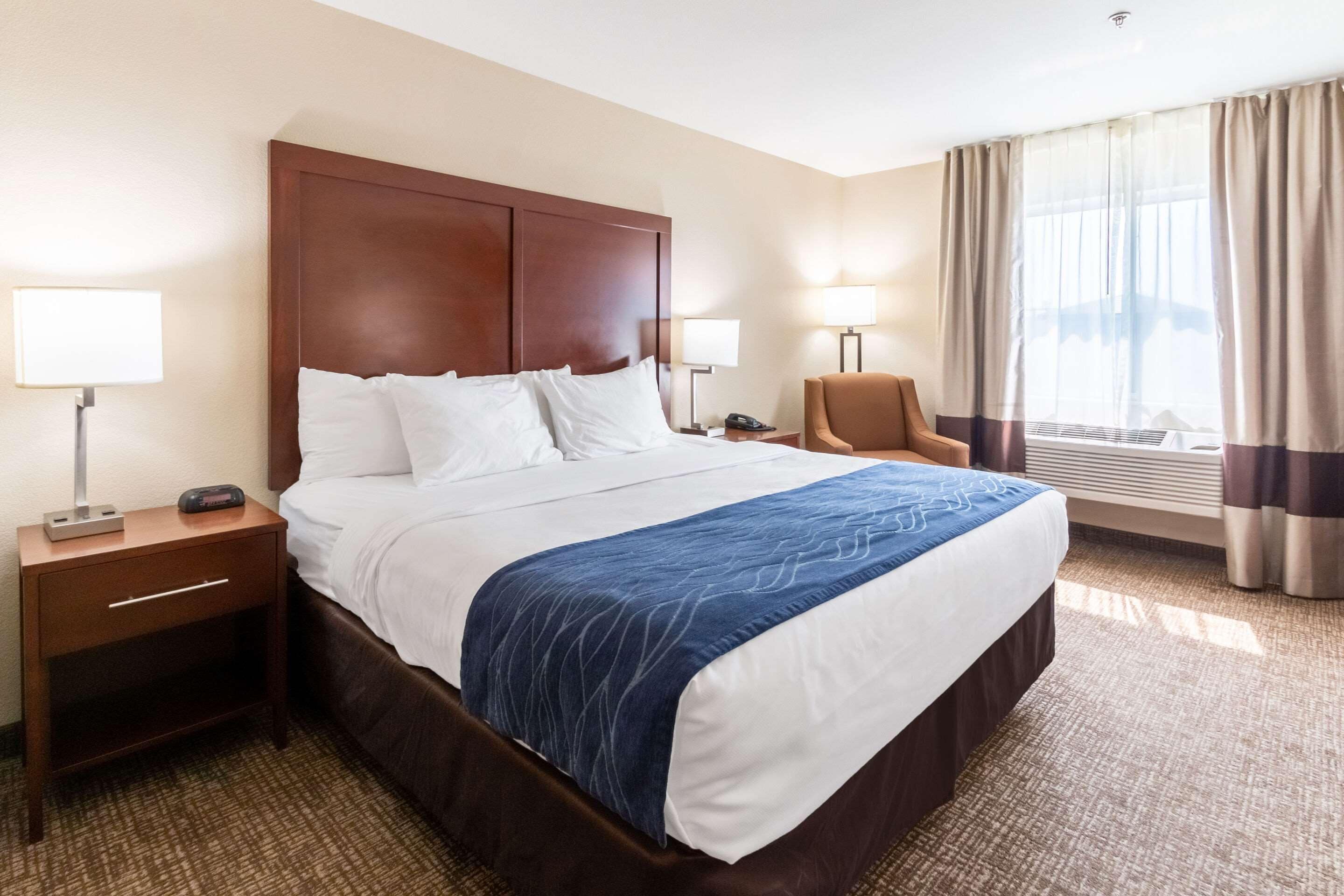 Habitación de hotel con una cama grande, dos mesitas de noche con lámparas, una silla y una ventana con cortinas.