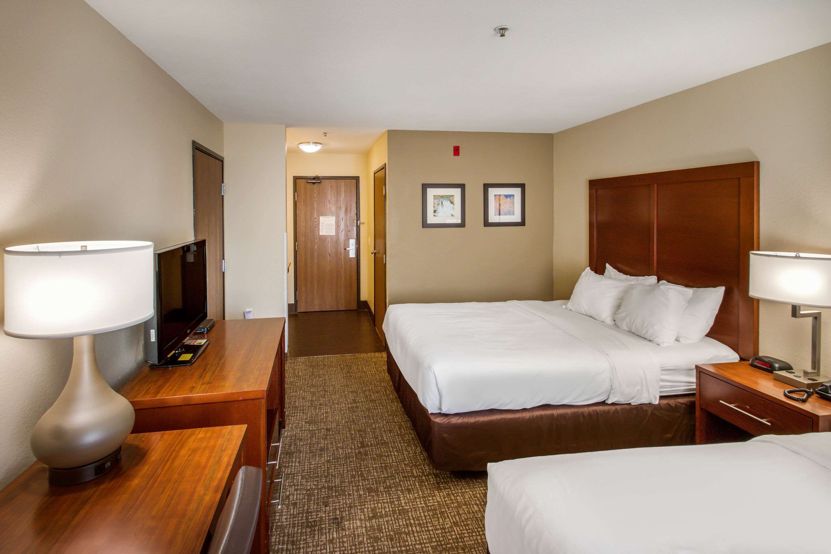 A hotel room with two beds, a desk, a chair, a TV, and a lamp.