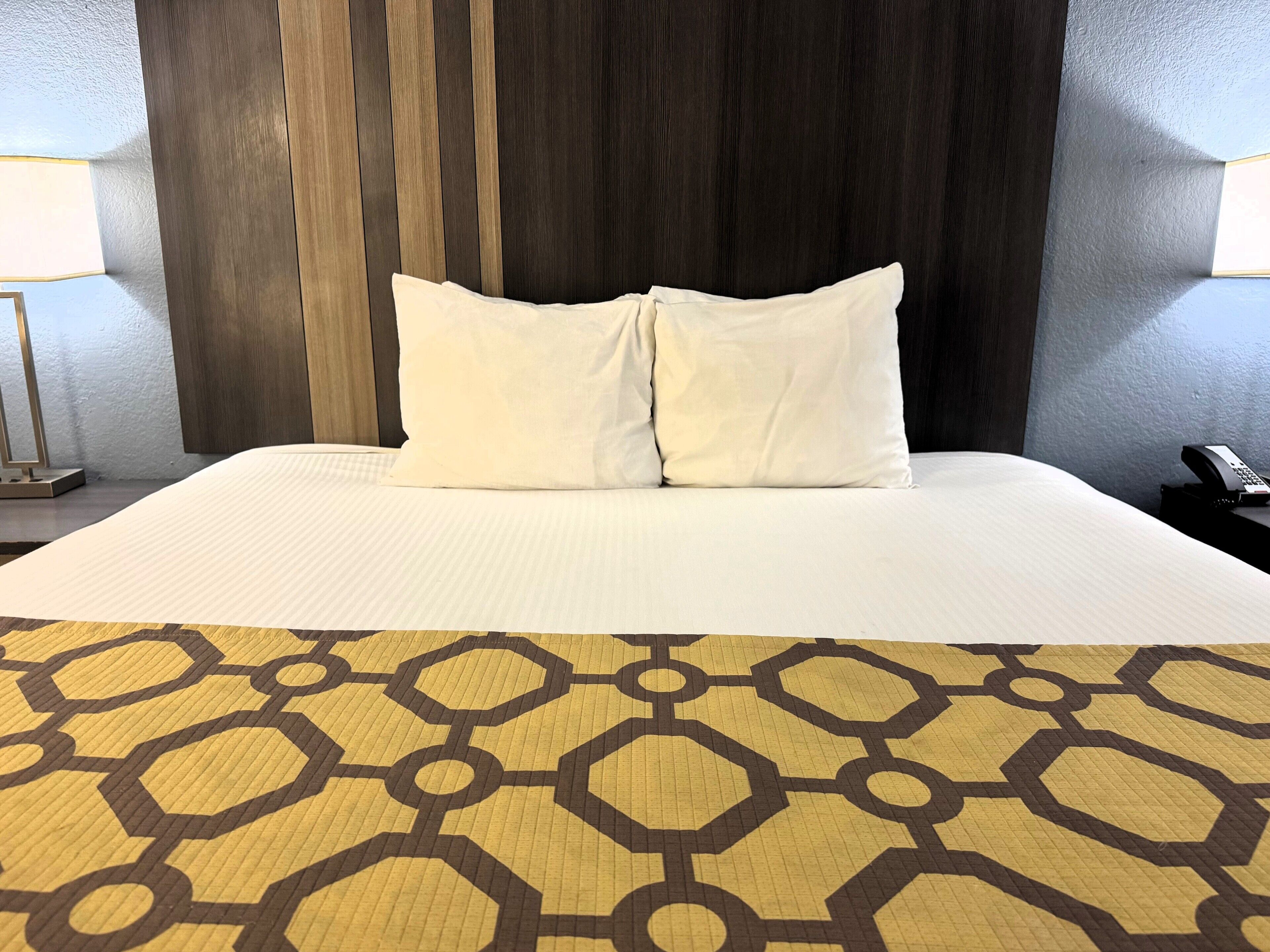 Une chambre d’hôtel avec un grand lit, deux oreillers, un tapis à motifs et une tête de lit en bois.